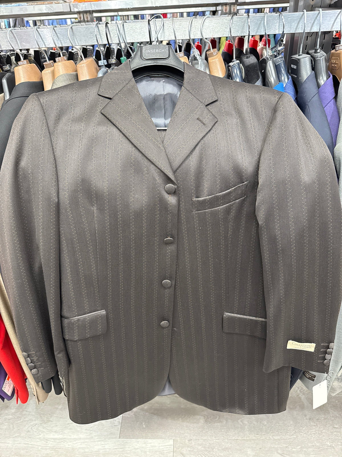 A Collezioni L’uomo Suit Brown jacket (40R ONLY) by Unique Design Menswear, featuring a dark pinstripe design with three buttons and three front pockets, is displayed on a hanger among other jackets in the store. Please note, this item is FINAL SALE with NO EXCHANGE or RETURN.