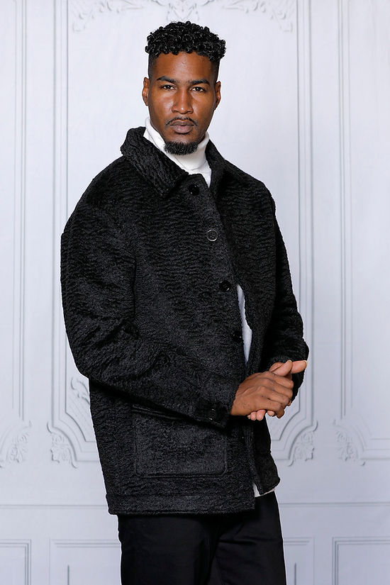 A man in a MANZINI CANADA Black Short Ripple Faux Fur Button Coat by Insomnia, tailored in regular fit, over a white turtleneck against a light gray backdrop.