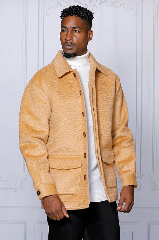 A man in a MANZINI CANADA Camel Short Ripple Faux Fur Button Coat by Insomnia, layered over a white turtleneck, stands against a light interior wall.