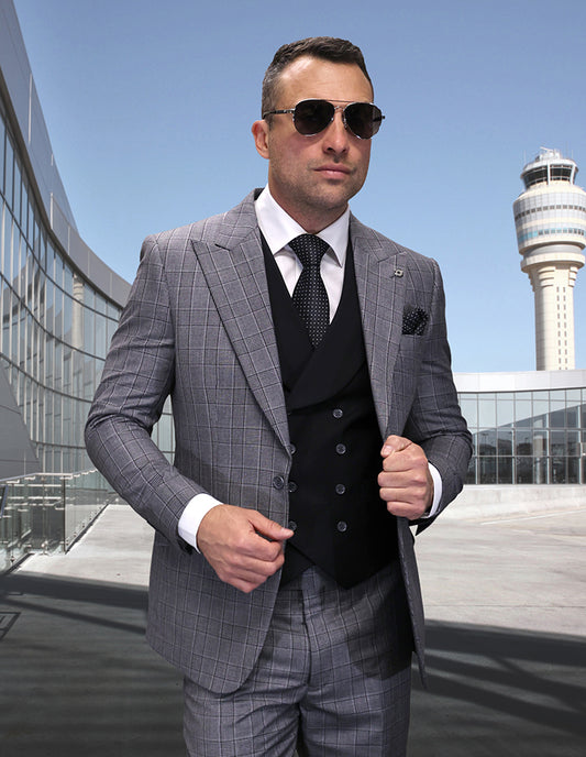 A man wearing the STATEMENT CLOTHING | CANON-BLACK suit by Statement Clothing, with sunglasses, stands outdoors near a modern building and control tower.