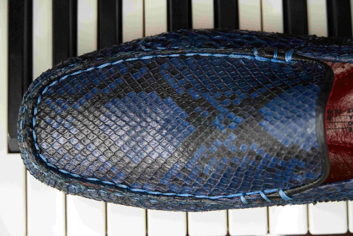 A pair of Capri Blue Jeans loafers by Marco Di Milano, crafted with a striking blue snakeskin texture; one shoe stands upright while the other lies on its side to reveal the black rubber sole—an elegant display of handcrafted luxury for those seeking exquisite exotic footwear.