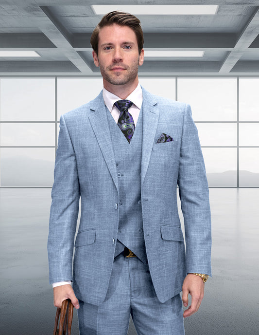 A man in the STATEMENT CLOTHING | CARNELI-BLUE suit by Statement Clothing stands in a modern, empty office with large windows, holding a brown briefcase. He pairs the light blue 3-piece suit with a white shirt and patterned tie.