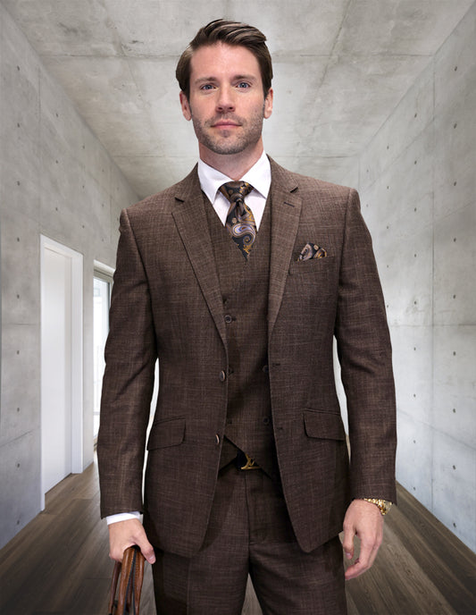 A man in the STATEMENT CLOTHING | CARNELI-BROWN modern fit Italian wool suit by Statement Clothing stands in a contemporary hallway with concrete walls and wooden floors, holding a leather bag.