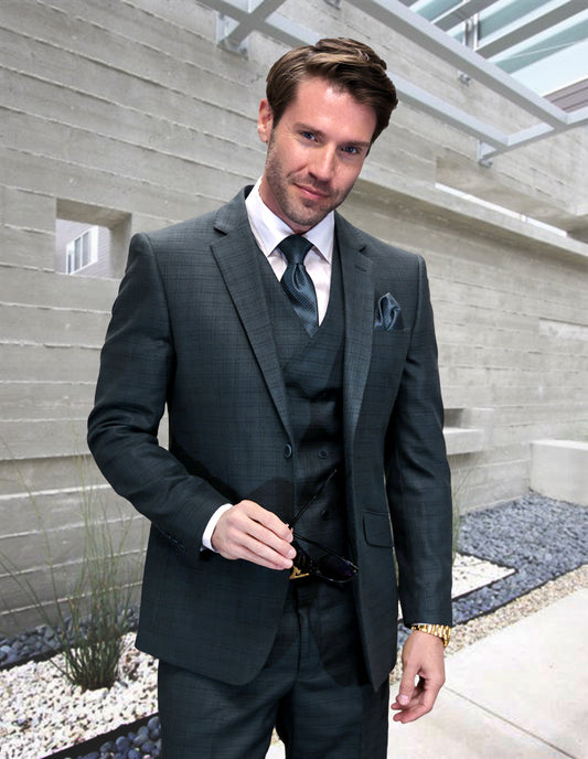 A man in a modern fit STATEMENT CLOTHING | CARNELI-HUNTER charcoal suit by Statement Clothing stands outdoors against a concrete wall, holding sunglasses and looking at the camera.