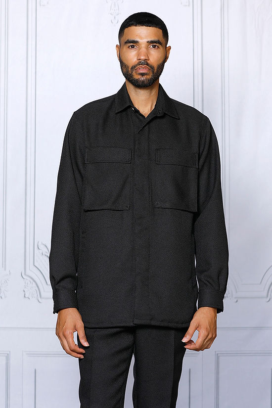A man faces forward in the Insomnia Cavalli 2PC Black Leisure Suit by Insomnia, featuring a black button-up shirt with front pockets and matching pants, set against a light-colored, ornate wall background.