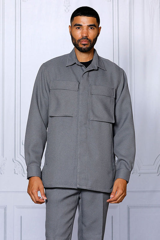 A man poses indoors against a light decorative wall, wearing the INSOMNIA CAVALLI 2PC Charcoal Leisure Suit by Insomnia—a matching poly-rayon blend suit with a textured button-up shirt with two pockets and coordinating grey pants.