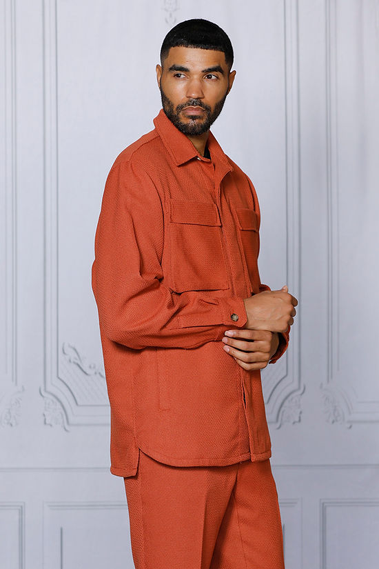 A man wearing the INSOMNIA CAVALLI 2PC Coral Leisure Suit by Insomnia, featuring a textured button-up shirt with chest pockets and matching pants, stands against a light-colored paneled wall.