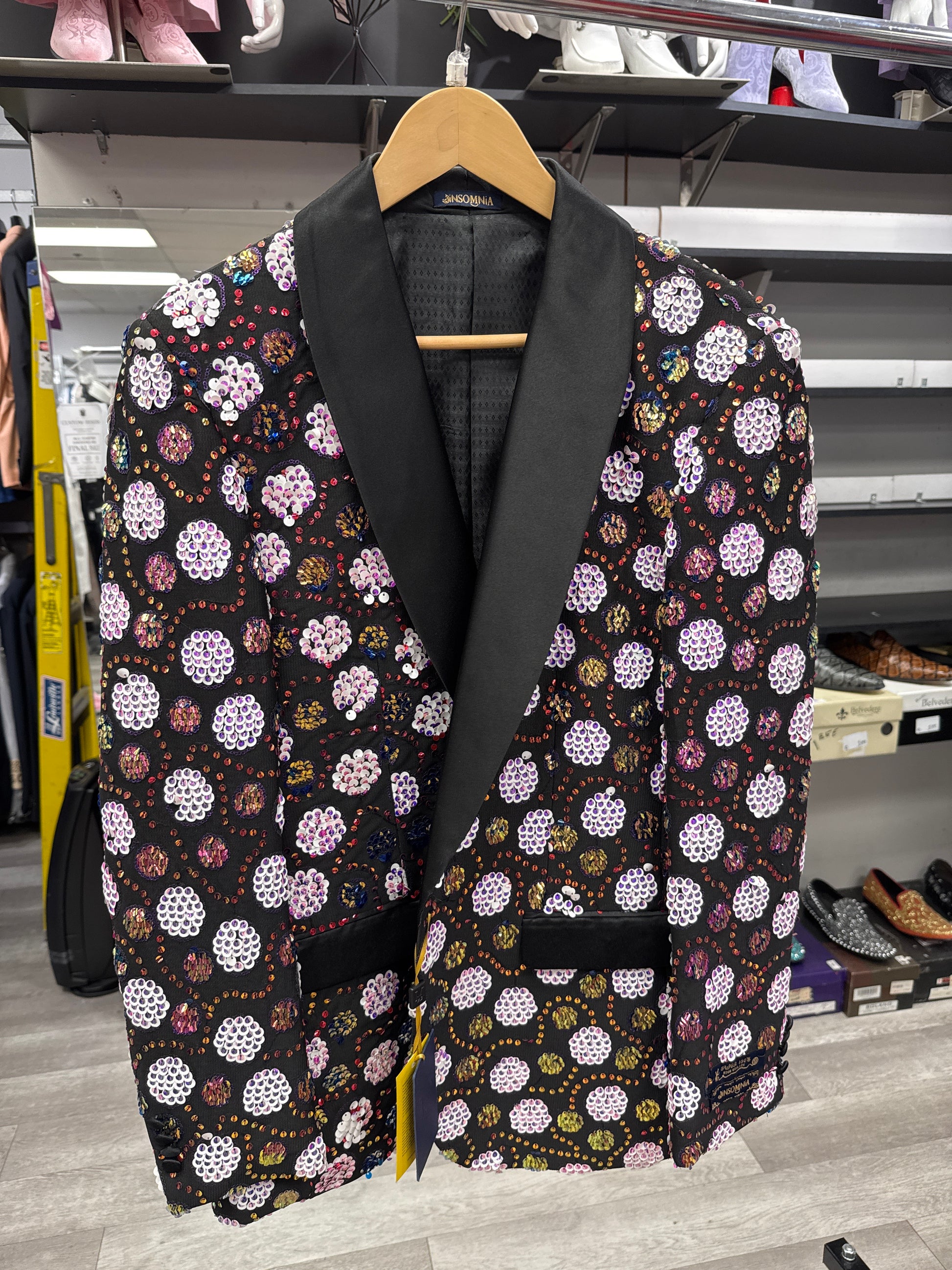 The EJ SAMUEL JACKET 1 CLEARANCE, featuring a black base with a vibrant floral pattern and black satin lapels, hangs on a wooden hanger in the clothing store. Please note, this item is final sale—no exchanges or returns.