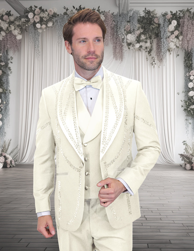 A man wears the STATEMENT CLOTHING | CHIC-OFF WHITE tuxedo by Statement Clothing, featuring sequin details, as he stands indoors against a backdrop of floral and draped curtains.