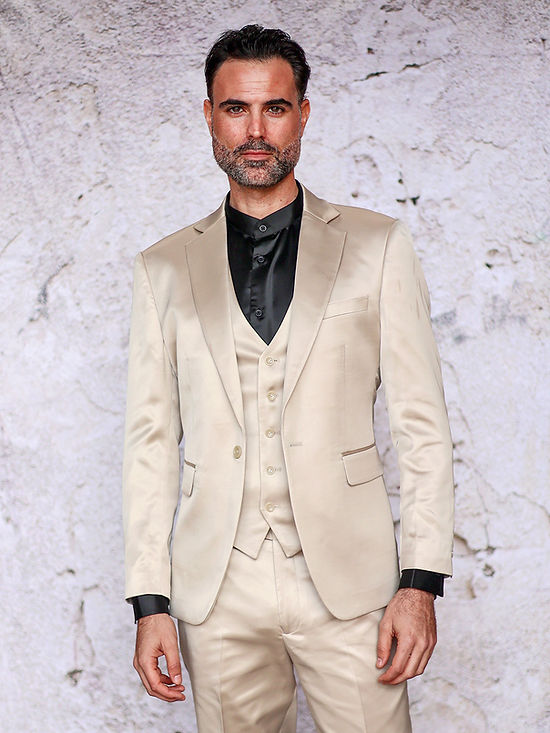 A man with short dark hair and a beard wears the Insomnia INSOMNIA CLASSICO 3PC Tailored Fit Satin Spandex Champagne Suit with a black shirt, standing before a textured light wall—ideal for a stylish night out.
