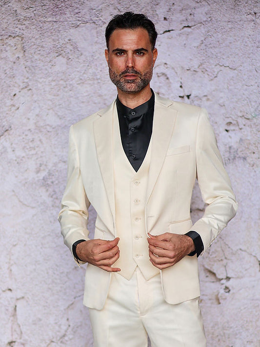 A man stands before a textured, light wall wearing the Insomnia INSOMNIA CLASSICO 3PC Tailored Fit Satin Spandex Cream Suit with a matching vest and slim fit black shirt.