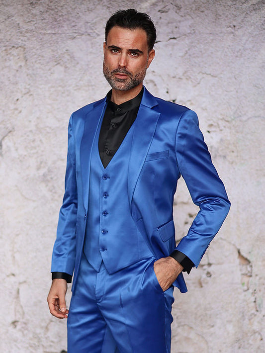 A man wears the INSOMNIA CLASSICO 3PC Tailored Fit Satin Spandex Royal Suit by Insomnia with a black shirt, standing against a textured light wall—ideal for a stylish night out.
