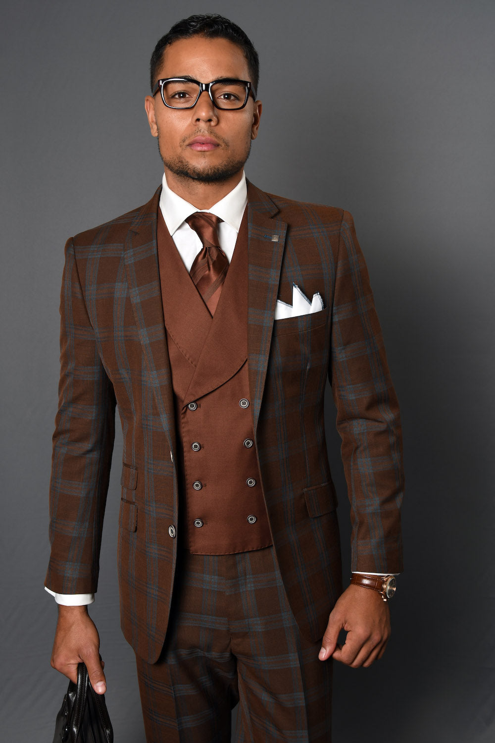 A man stands against a gray background, wearing the STATEMENT CLOTHING | COMO-COPPER plaid copper suit by Statement Clothing with matching vest, brown tie, white shirt, pocket square, and holding a black bag.