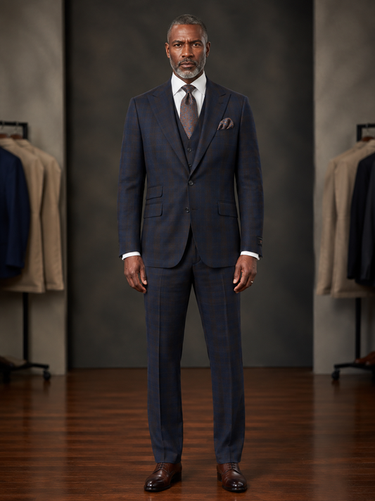 A man stands in a showroom wearing the Tiglio Rosso Orvietto Navy & Brown Windowpane Semi Wide Leg TL7506 suit, paired with a crisp white shirt, coordinating tie, and brown dress shoes. Clothing racks filled with jackets are visible in the background, emphasizing the sophisticated appeal of Tiglio Rosso three piece suits.