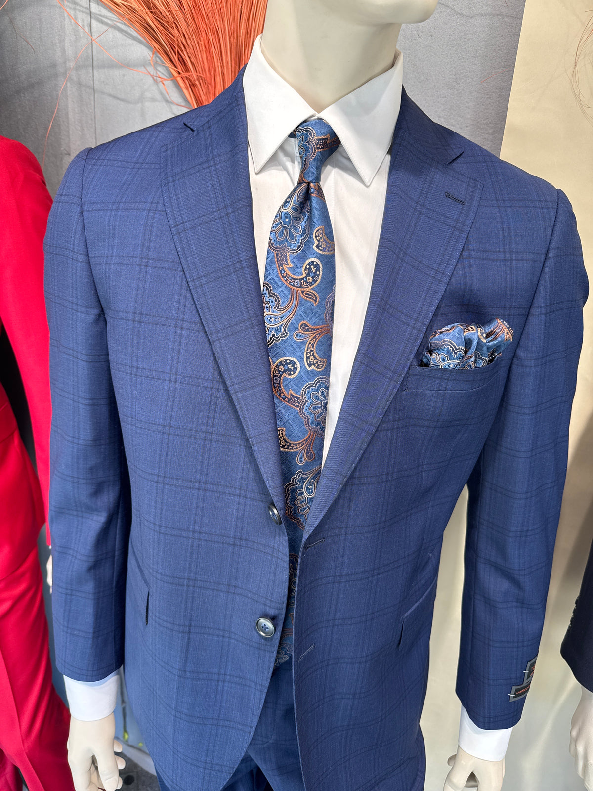 A man stands facing forward in a Tiglio Luxe Dolcetto Modern Fit Wool Suit CB3508 CR3508, complemented by a white shirt, patterned tie, and brown shoes; clothing racks and a neutral background appear behind him.