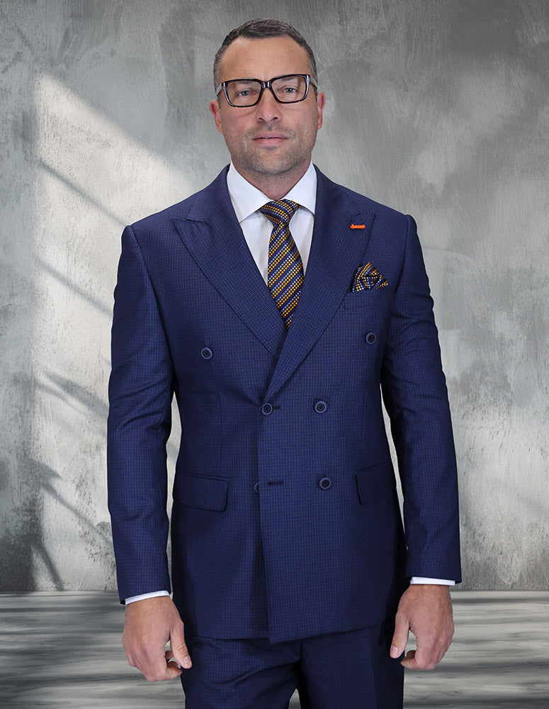 A man in the STATEMENT CLOTHING | DB-SALOTE-BLUE modern fit Italian wool double-breasted suit by Statement Clothing, white shirt, patterned tie, and glasses stands against a textured gray background.
