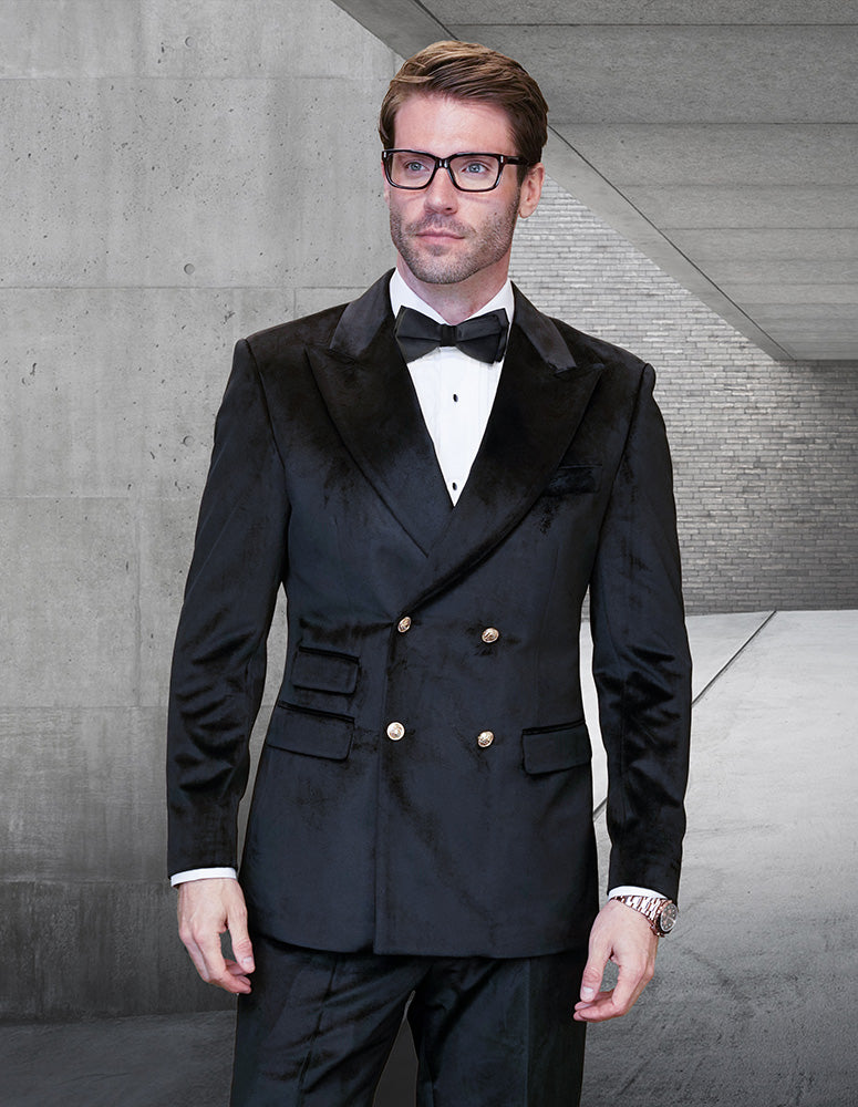 A man in the STATEMENT CLOTHING | DB-VELVET-BLACK double-breasted suit by Statement Clothing, with a white shirt, black bow tie, and glasses, stands in a modern concrete hallway.