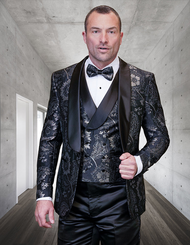 A man wears Statement Clothing’s DELANO-3-BLACK, a modern fit black floral shawl lapel tuxedo with a bow tie, standing in a sleek hallway with concrete walls and dark wood floors.