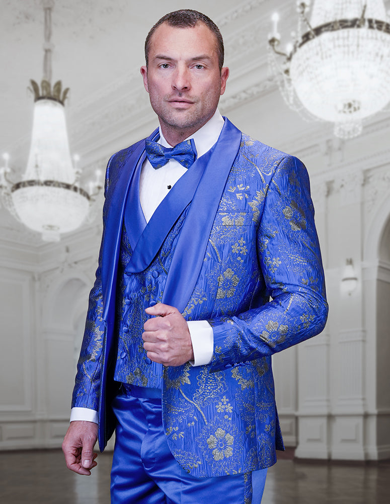 A man stands in an elegant, chandelier-lit ballroom wearing the STATEMENT CLOTHING | DELANO-3-ROYAL tuxedo by Statement Clothing, featuring a royal blue and gold floral patterned shawl lapel and a bow tie.