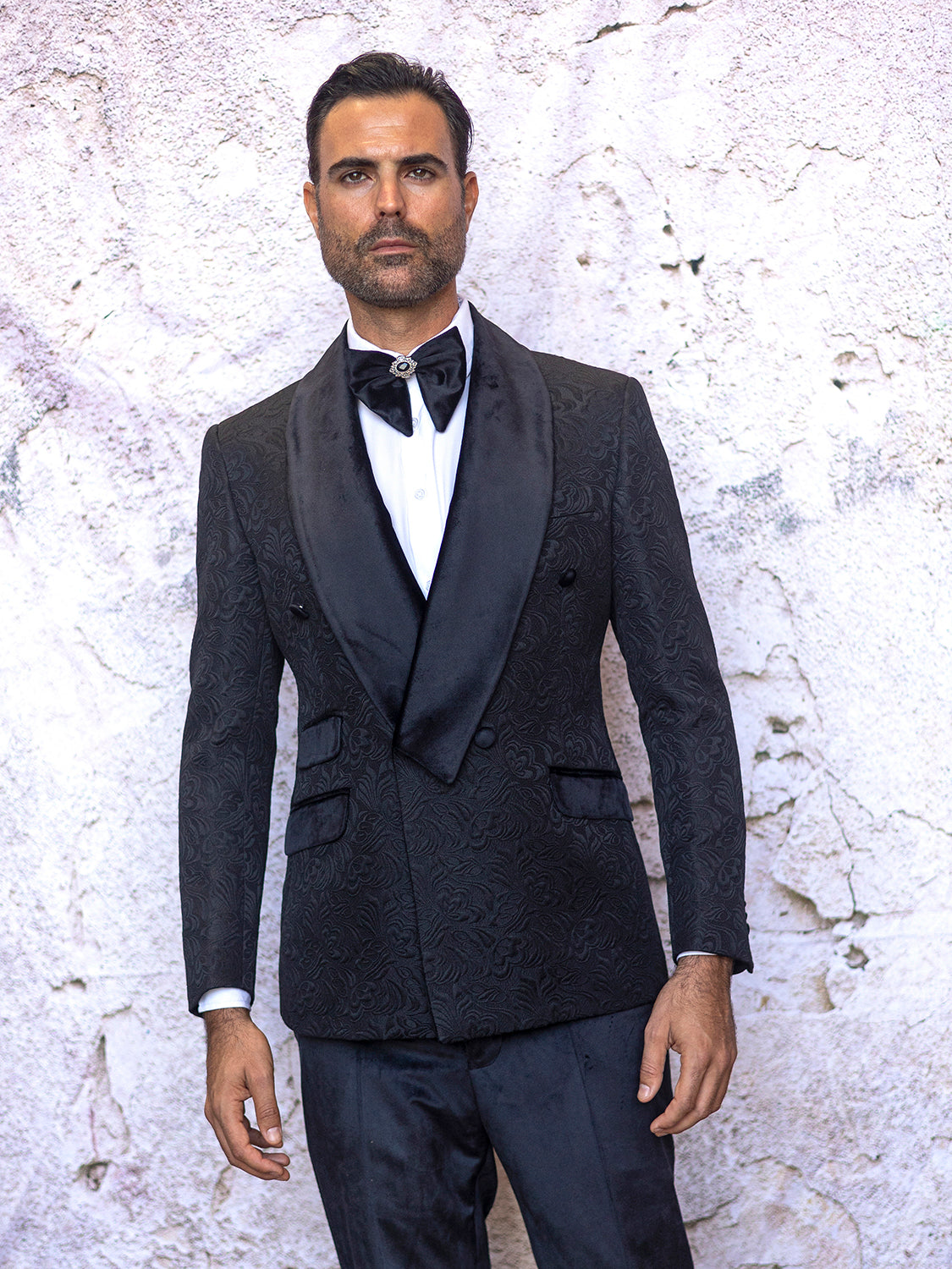 A man in an Insomnia MANZINI DELANO 2-Piece Slim Fit Double Breast Floral Texture Black Tuxedo, featuring a white shirt and velvet-trimmed bow tie, stands against a textured white wall.