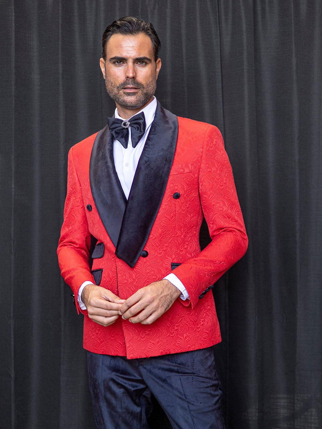 A man wearing an Insomnia MANZINI DELANO 2-Piece Slim Fit Double Breast Floral Texture Red Tuxedo stands in front of a black curtain, paired with a bow tie and black velvet trim.