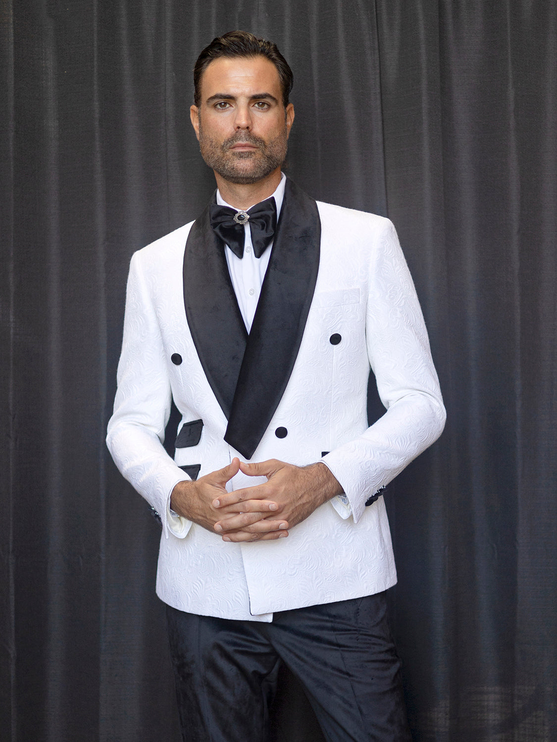 An individual wearing an Insomnia MANZINI DELANO 2-Piece Slim Fit Double Breast Floral Texture White Tuxedo, featuring a black bow tie and velvet-trimmed lapels, stands against a dark curtain backdrop with his hands clasped.