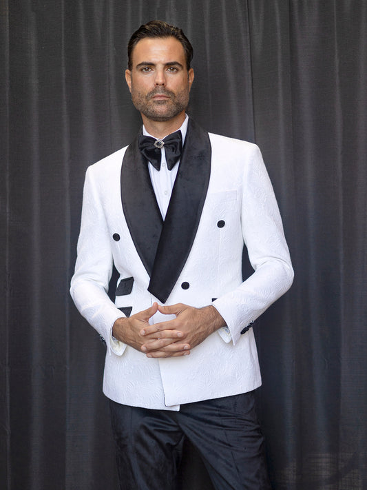 An individual wearing an Insomnia MANZINI DELANO 2-Piece Slim Fit Double Breast Floral Texture White Tuxedo, featuring a black bow tie and velvet-trimmed lapels, stands against a dark curtain backdrop with his hands clasped.