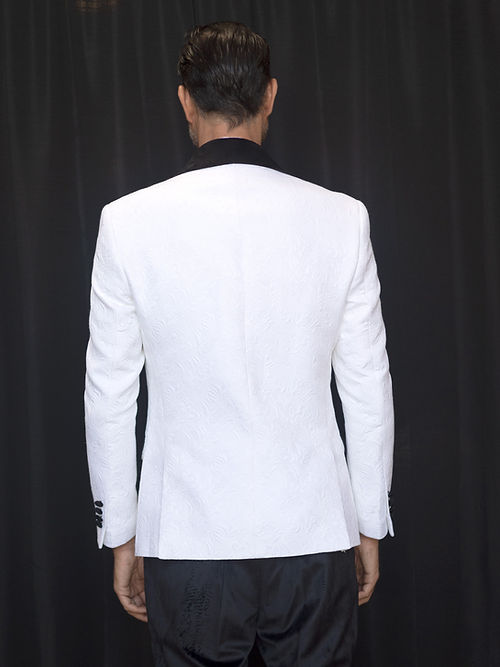 An individual wearing an Insomnia MANZINI DELANO 2-Piece Slim Fit Double Breast Floral Texture White Tuxedo, featuring a black bow tie and velvet-trimmed lapels, stands against a dark curtain backdrop with his hands clasped.