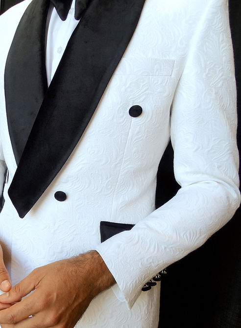 An individual wearing an Insomnia MANZINI DELANO 2-Piece Slim Fit Double Breast Floral Texture White Tuxedo, featuring a black bow tie and velvet-trimmed lapels, stands against a dark curtain backdrop with his hands clasped.