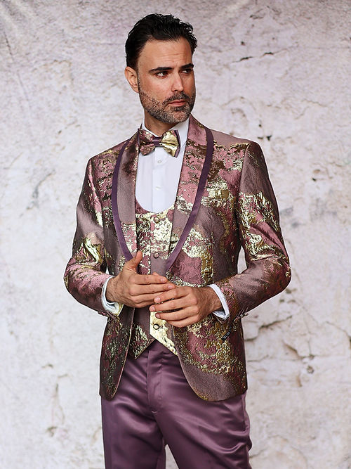 A man in a slim fit maroon MANZINI DELMAS tuxedo from Insomnia, featuring a shiny metallic pattern with a matching bow tie and vest, stands elegantly in front of a textured wall, looking to the side.
