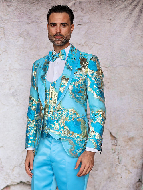 A man in the Insomnia MANZINI DELMAS 3-Piece Slim Fit Shiny Metallic Pattern Sky Tuxedo Prom Suit, featuring a vibrant blue and gold floral design, with a coordinating bow tie, stands against a textured backdrop.