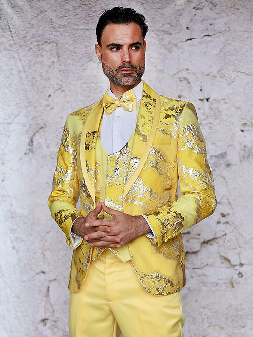 A man in an Insomnia MANZINI DELMAS 3-piece slim fit yellow tuxedo with a bow tie stands against a textured wall, his hands clasped. The suit's shiny metallic jacquard pattern enhances his elegant appearance.