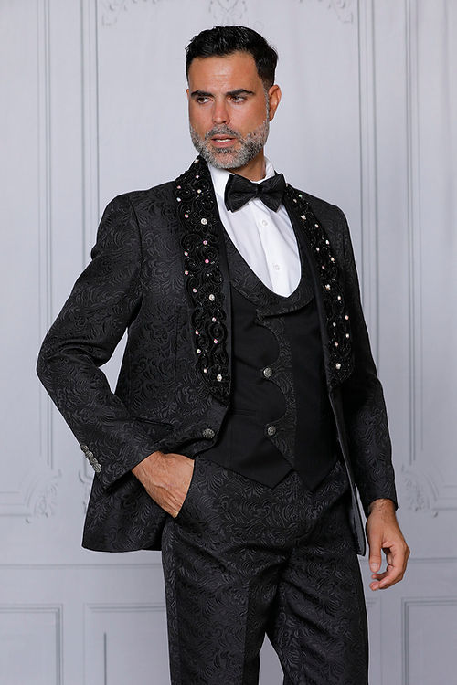 A man in an Insomnia MANZINI DIOR 3-Piece Slim Fit Rhinestone & Bead Trim Floral Black Tuxedo Suit with embellished lapels, elegantly wearing a bow tie, stands against a light-colored background.