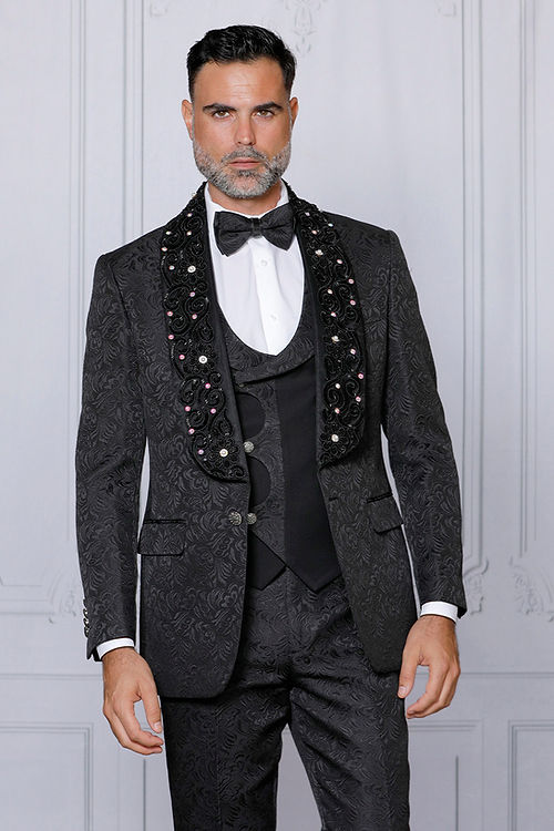 A man in an Insomnia MANZINI DIOR 3-Piece Slim Fit Rhinestone & Bead Trim Floral Black Tuxedo Suit with embellished lapels, elegantly wearing a bow tie, stands against a light-colored background.