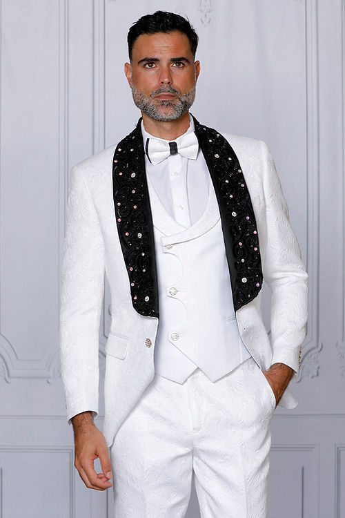 Against a light wall, a confident man stands in an Insomnia MANZINI DIOR 3-piece slim fit rhinestone & bead trim floral white tuxedo suit with a black embellished scarf.