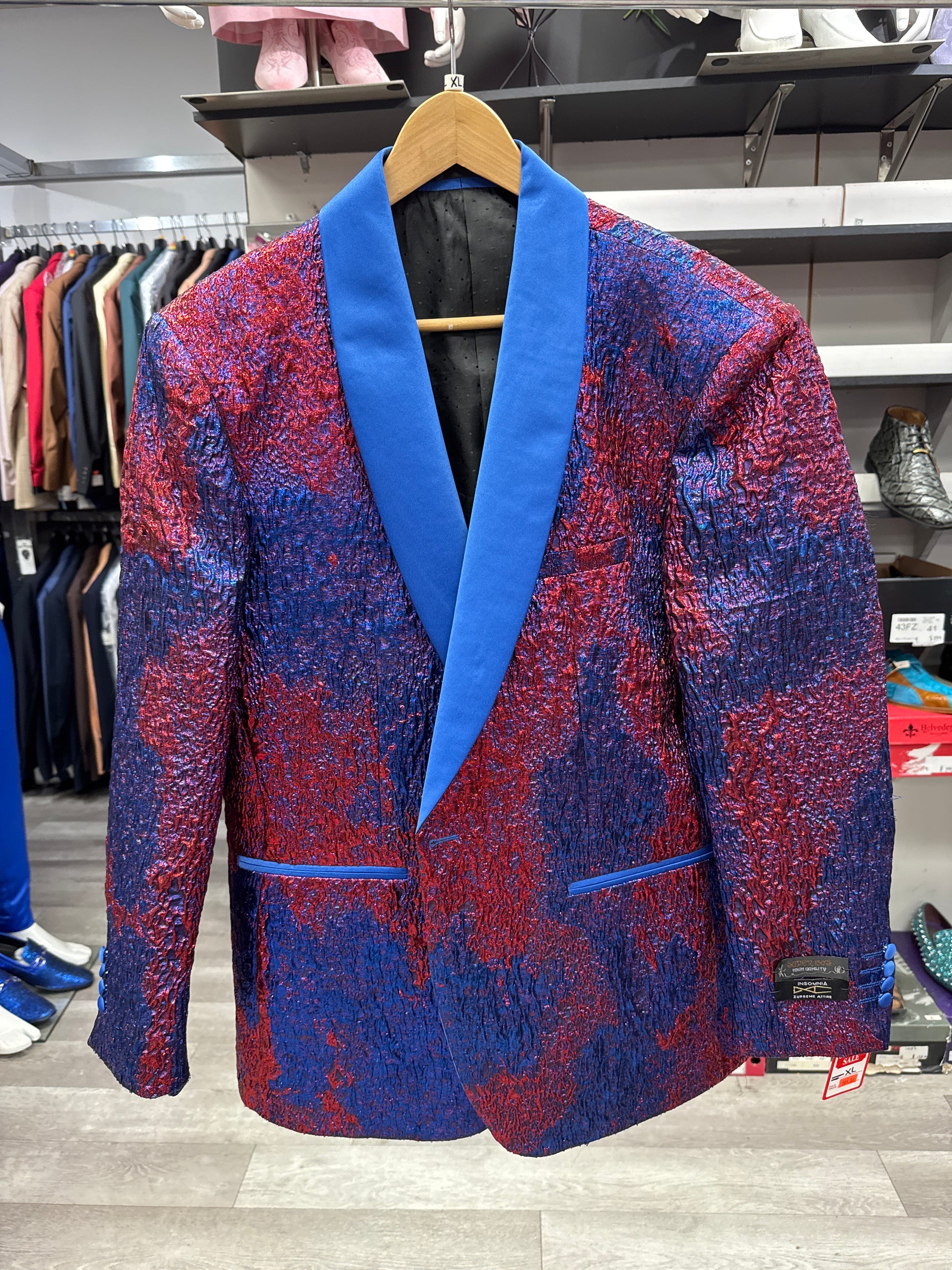 The EJ SAMUEL JACKET 3 CLEARANCE, featuring a red and blue crinkled pattern and a vibrant blue shawl collar, is displayed on a wooden hanger in the clothing store. Please note that this item is final sale—no returns or exchanges will be accepted.