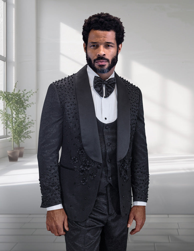 A man stands indoors wearing the STATEMENT CLOTHING | ELIO-BLACK textured modern fit tuxedo by Statement Clothing, complete with a bow tie and vest. A potted plant and large windows are visible in the background.