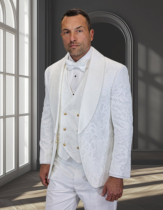 A man wears the STATEMENT CLOTHING | EMPORIO-IVORY slim fit white tuxedo by Statement Clothing, featuring a patterned shawl lapel, white bow tie, and double-breasted velvet vest in a room with arched windows and grey walls.