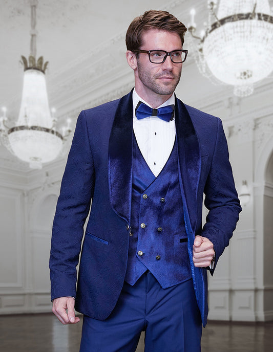 A man wearing the STATEMENT CLOTHING | EMPORIO-SAPPHIRE slim fit blue velvet tuxedo by Statement Clothing, featuring a shawl lapel and bow tie, stands in an elegant, chandelier-lit ballroom.