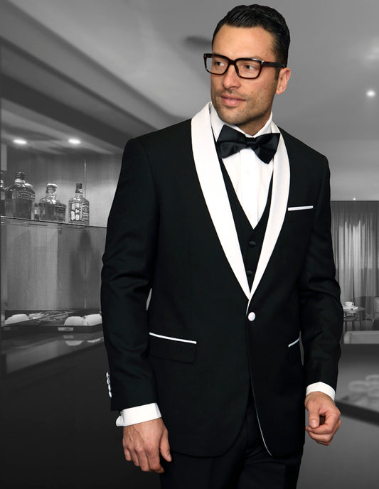 A man models the STATEMENT CLOTHING | ENCORE-BLACK tuxedo by Statement Clothing, featuring white lapels and a bow tie, tailored for a sleek fit as he stands in a modern, dimly lit room with a bar behind him.