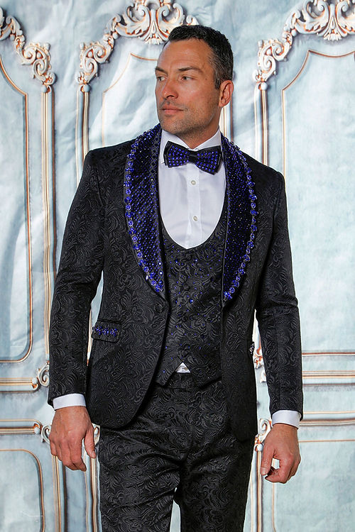 A man stands out in an Insomnia Manzini Enzo 3-piece slim fit suit featuring a woven pattern in black and royal with a rhinestone embellishment, completed by a blue sequined lapel and bow tie, set against an ornate backdrop.