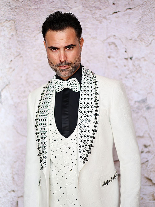A man with short dark hair wears an Insomnia MANZINI ENZO 3-Piece Slim Fit Fancy Rhinestone Woven Pattern White/Black Suit and a polka dot bow tie, standing against a light stone wall.