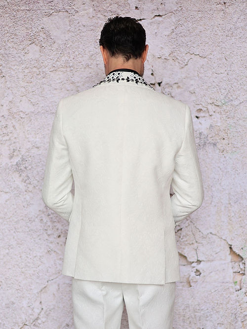 A man with short dark hair wears an Insomnia MANZINI ENZO 3-Piece Slim Fit Fancy Rhinestone Woven Pattern White/Black Suit and a polka dot bow tie, standing against a light stone wall.