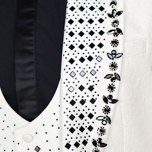 A man with short dark hair wears an Insomnia MANZINI ENZO 3-Piece Slim Fit Fancy Rhinestone Woven Pattern White/Black Suit and a polka dot bow tie, standing against a light stone wall.