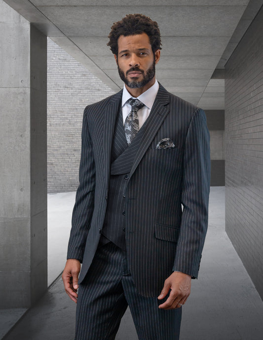 A man with a beard and short curly hair wears the STATEMENT CLOTHING | EXPLO-BLACK suit by Statement Clothing, paired with a white shirt and patterned tie, as he stands in a sleek concrete hallway.