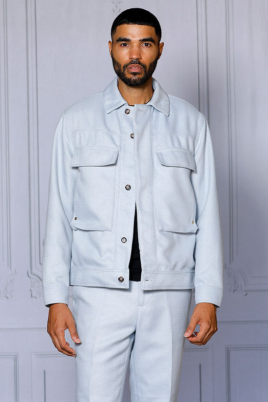 A man wearing the INSOMNIA FENDI 2PC Silver Leisure Suit by Insomnia stands against a decorative light-colored wall, looking to his left.