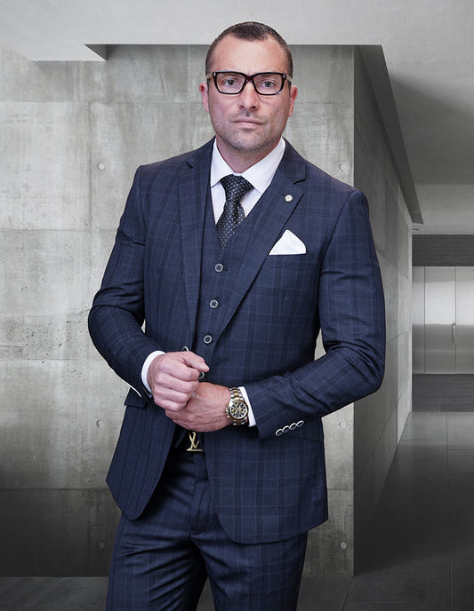 A man wearing the STATEMENT CLOTHING | FERETI-CHARCOAL suit by Statement Clothing, paired with a white pocket square and glasses, stands in a minimalist hallway with concrete walls.