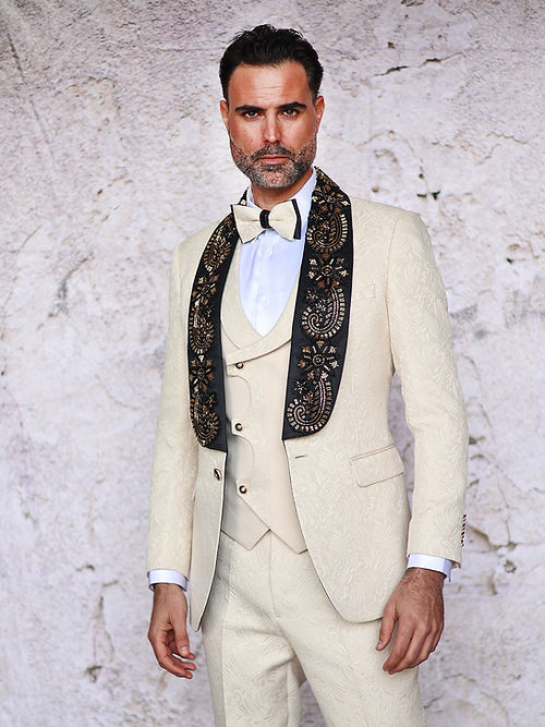 A man in a Manzini Giorgio 3PC slim fit tan tuxedo with a floral pattern, black bead lapel by Insomnia, stands against a textured stone wall, wearing a white shirt and bow tie.