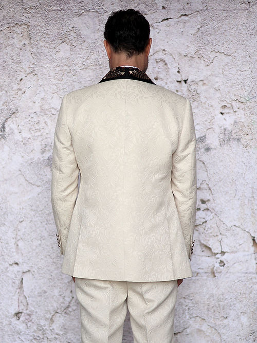 A man in a Manzini Giorgio 3PC slim fit tan tuxedo with a floral pattern, black bead lapel by Insomnia, stands against a textured stone wall, wearing a white shirt and bow tie.