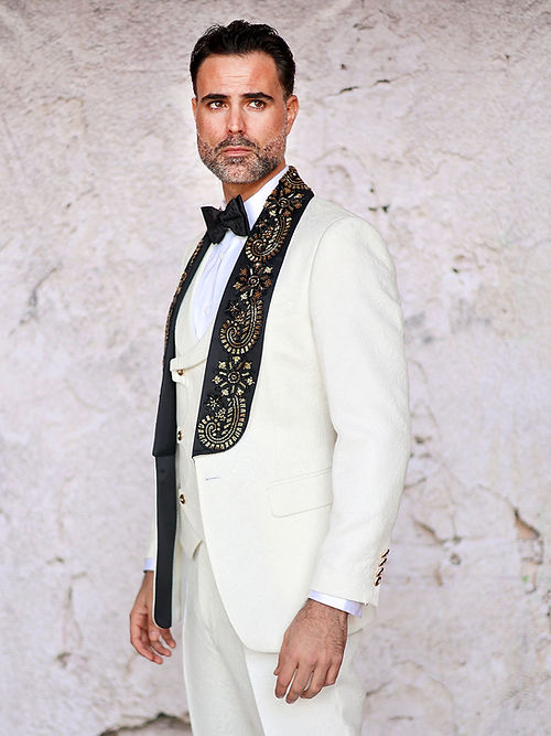 A man in an Insomnia MANZINI GIORGIO 3PC Slim Fit Fancy Floral Pattern White Tuxedo with a bead lapel stands against a textured light-colored wall.